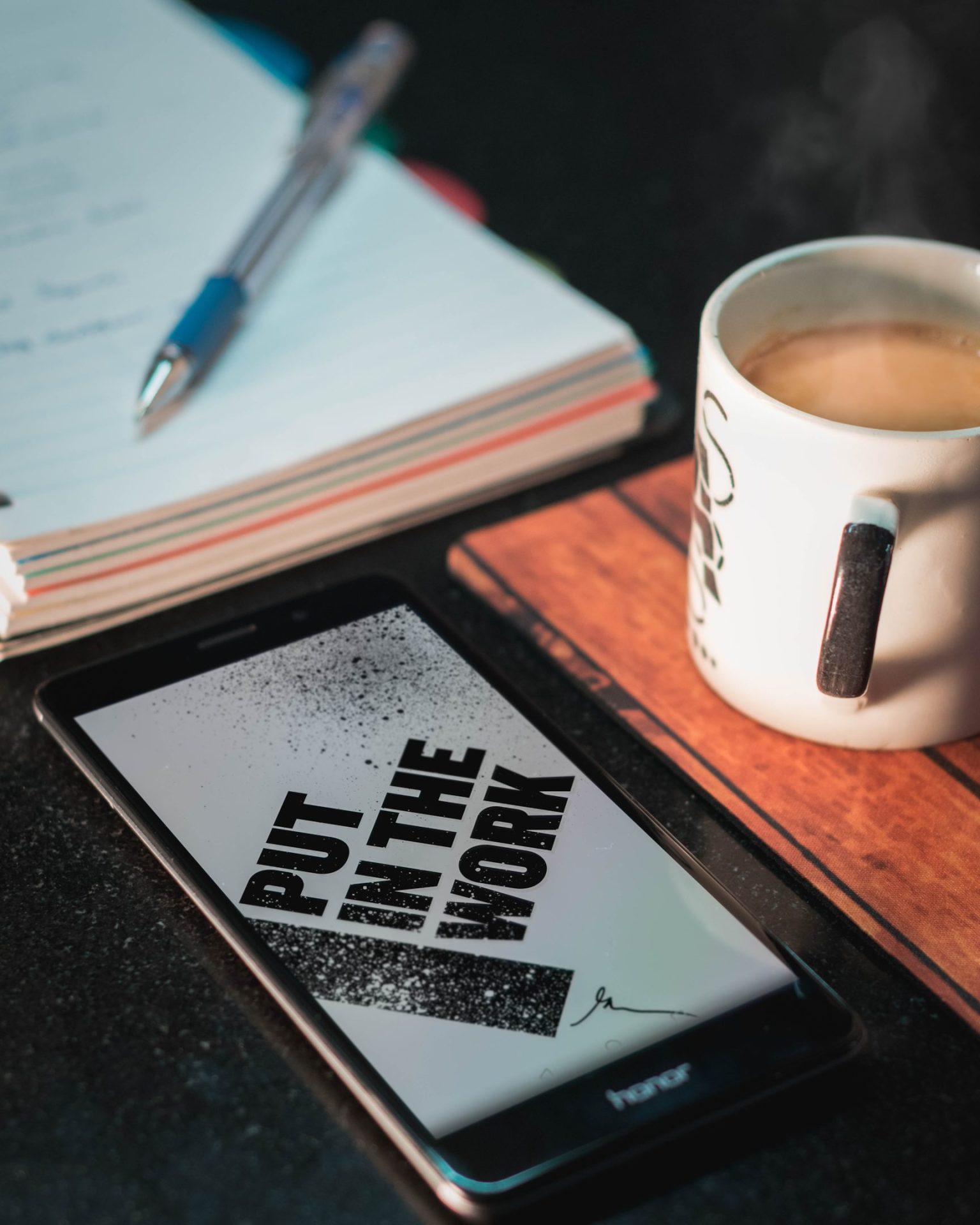 photo of phone on the desk together with a cup of coffee, a notebook, and a pen